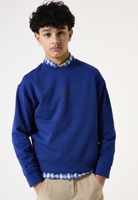 Sudadera azul con cuello redondo y puños de canalé, que presenta texto en la parte delantera. Llevada sobre una camisa de cuadros azul y blanca con cuello.