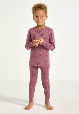 Nuori kiharatukkainen lapsi hymyilee, yllään yhteensopivat liilat yöpuku, joissa on eläin- ja luontokuvioita, seisomassa paljain jaloin valkoista taustaa vasten.