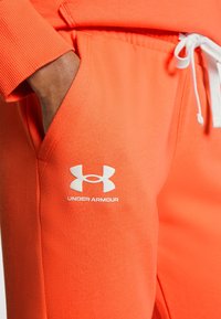 Orange joggerbyxor med mjuk textur, som har en vit dragsko och en Under Armour-logotyp på vänster lår. Fickor ingår.