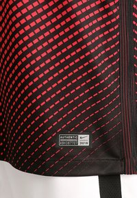 Tissu athlétique noir avec des rayures diagonales rouges et un motif texturé, présentant une étiquette d'authenticité dans le coin inférieur droit.