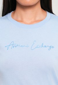 Lichtblauwe katoenen T-shirt met een ronde halslijn en glinsterende blauwe tekst "Armani Exchange" op de voorkant. Gladde stofstructuur.