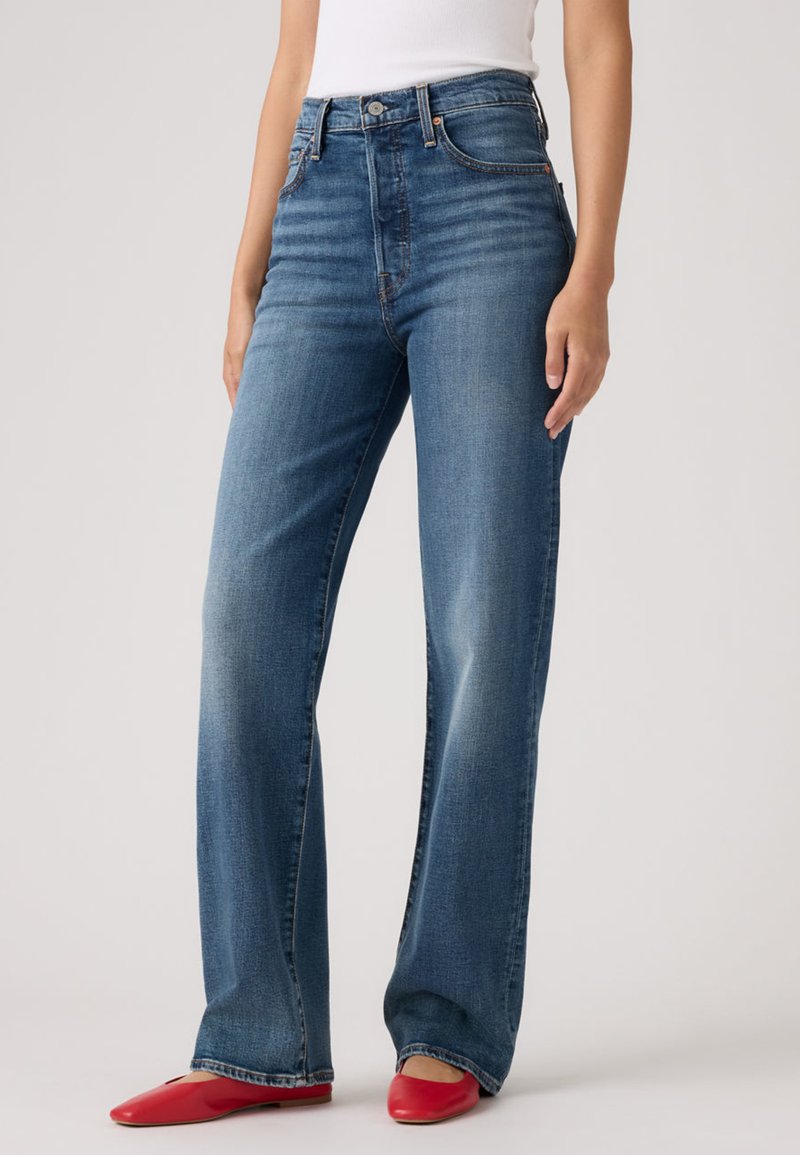 Γυναίκα που φοράει μπλε τζιν με ψηλή μέση και ίσια μπατζάκια, συνδυασμένα με κόκκινα μπαλαρίνια και λευκό αμάνικο τοπ, στέκεται μπροστά σε απλό φόντο.