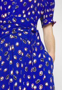 Blue fabric dress featuring a scattered pattern of black, white, and orange shapes, with a tied waist and pockets. Short sleeves included.