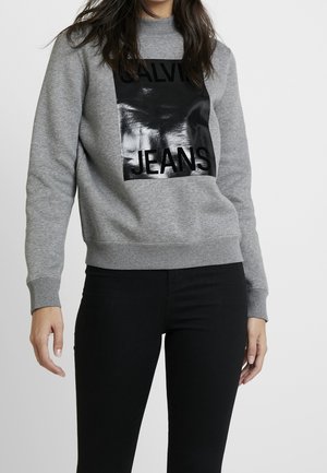 Femme portant un sweat-shirt gris avec un carré noir brillant et les mots "CALVIN JEANS" imprimés sur le devant et un pantalon noir.
