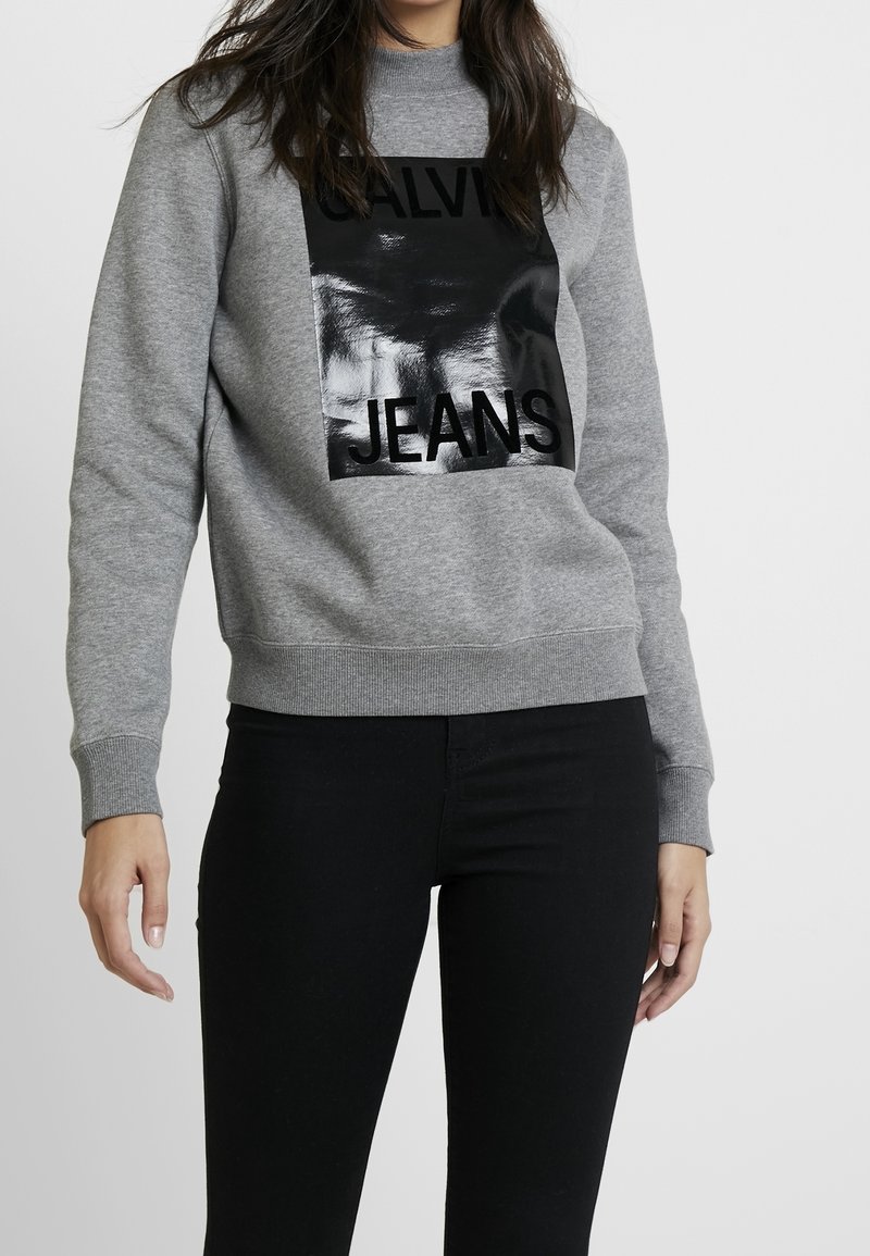 Femme portant un sweat-shirt gris avec un carré noir brillant et les mots "CALVIN JEANS" imprimés sur le devant et un pantalon noir.