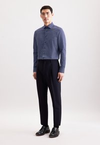 Uomo in camicia button-up a righe blu navy, pantaloni neri su misura e scarpe eleganti nere, in piedi su uno sfondo chiaro semplice.