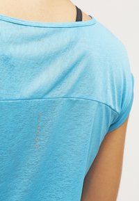 Světle modré sportovní tričko s krátkým rukávem a hladkou texturou. Má jemný horizontální šev a šest dekorativních kulatých akcentů na zádech.
