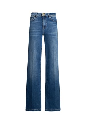 Frau trägt hoch taillierte, weit geschnittene blaue Jeans mit silberner Gürtelschnalle, weißes ärmelloses Oberteil und weiße spitze Schuhe.