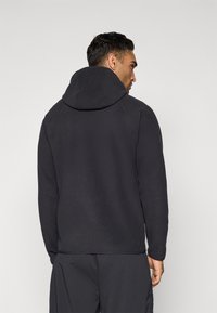 Man wearing a black hooded fleece jacket and black pants, viewed from behind against a plain light background.