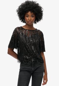 Zwart cropped top met korte mouwen, gemaakt van doorschijnende stof. Versierd met pailletten die franjes vormen. Gecombineerd met zwarte spijkerbroek.