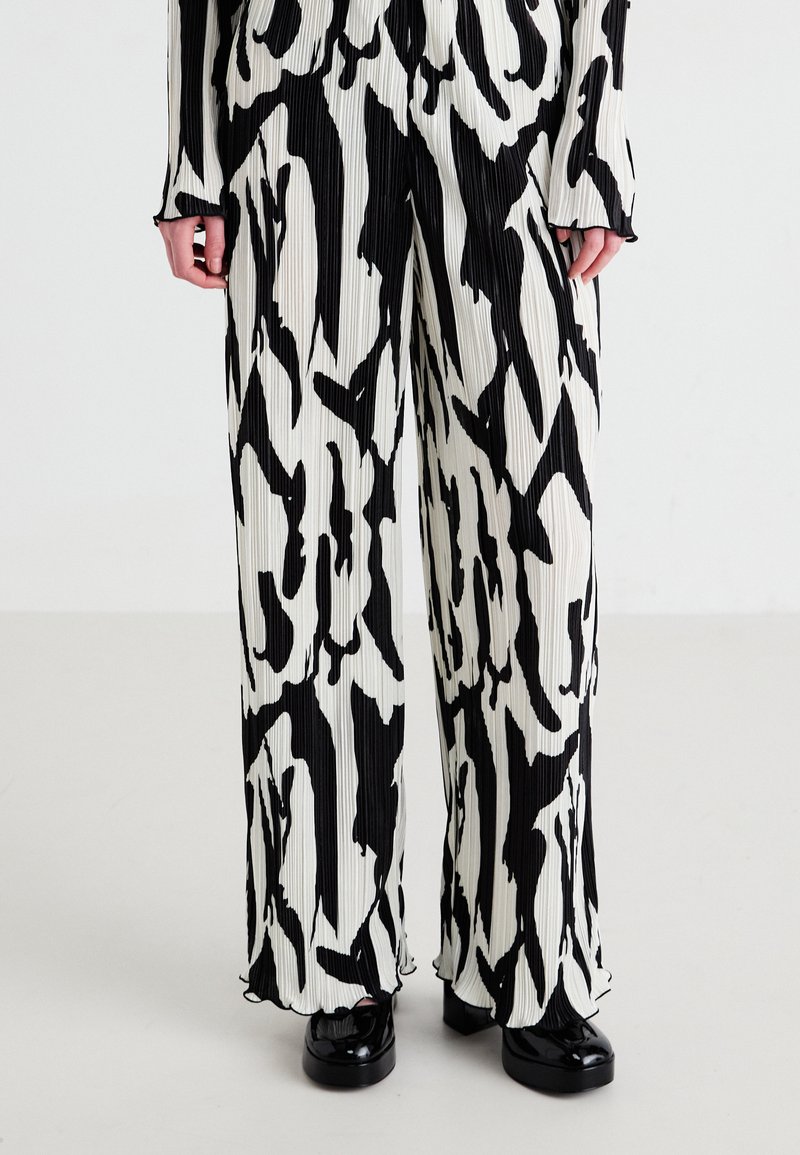 Pantalons plissés à motif noir et blanc, avec une coupe ample, présentant une texture lisse et des ourlets bien finis, associés à des chaussures noires brillantes.