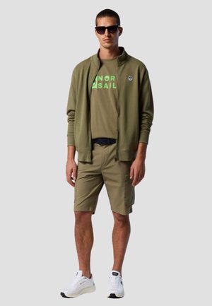 Giacca zip in verde oliva sopra una maglietta coordinata con stampa verde, abbinata a pantaloni corti beige e sneakers bianche. Modello con occhiali da sole.