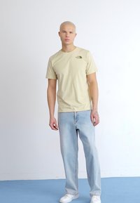 Camiseta amarilla clara de manga corta con un pequeño logo verde, combinada con jeans desgastados azul claro y zapatillas blancas.
