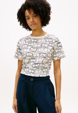 Femme aux cheveux bouclés portant une chemise blanche à manches courtes avec des lignes abstraites noires et un pantalon bleu marine taille haute avec une ceinture.