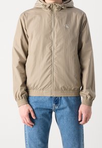 Béžová lehká bunda s kapucí, elastickými manžetami a pasem, zipovým zapínáním a malým logem na levé straně hrudi.