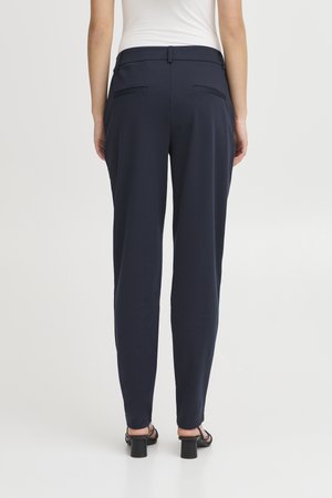 Naine, kes kannab tumesiniseid kohandatud pükse ja musti kontsaga sandaale, seisab valge ühevärvilise tausta ees, seljaga kaamera poole.