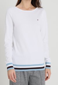 Witte lange mouwen shirt met een ronde halslijn, met gestreepte manchetten in blauw, zwart en wit, en een klein logo op de borst.