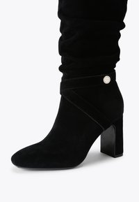 Bottine en suède noir avec un bout pointu, texture douce, détail de fronces et un talon bloc. Présente une sangle décorative et un matériel rond.
