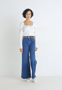 Top bianco a maniche lunghe con dettagli in pizzo, abbinato a jeans blu a gamba larga a vita alta. La modella indossa sneakers bianche e una cintura marrone.