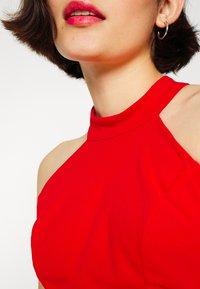 Top rouge à col licou au design épuré, avec des épaules découvertes et une texture lisse. Mise en valeur par un décolleté minimaliste.