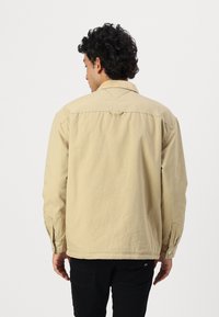 Beige jakke med en afslappet pasform, knapmanchetter og en detaljeret rygklap. Stoffet ser tekstureret og en smule rynket ud.