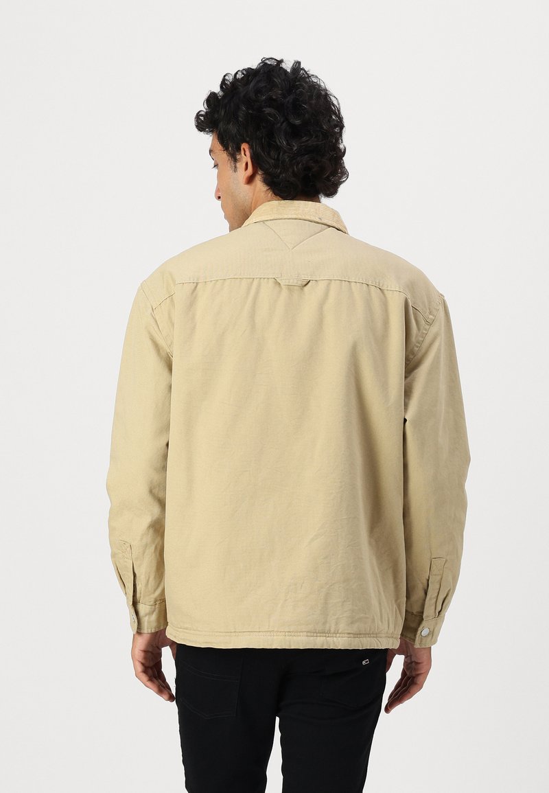 Chaqueta beige con un corte relajado, puños abotonados y un detalle de yugo en la parte posterior. La tela parece texturizada y ligeramente arrugada.