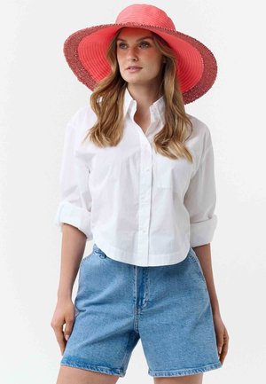 Mujer con un sombrero de ala ancha color coral, camisa blanca de botones con las mangas remangadas y shorts de mezclilla azul de tiro alto.