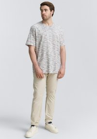 T-shirt à manches courtes, gris clair avec un motif texturé et ondulé ; associé à un pantalon couleur crème et des baskets blanches. Design simple.
