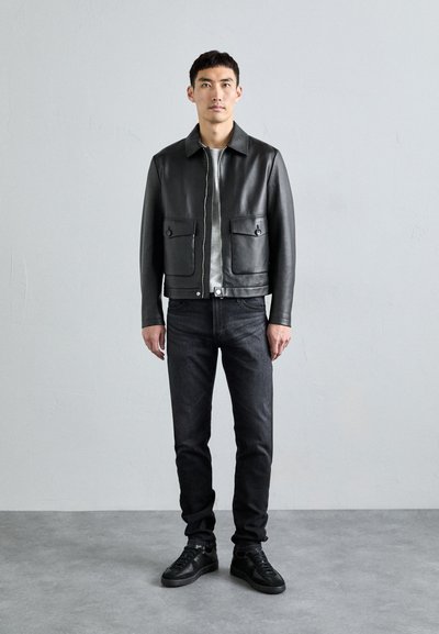 Svart läderjacka med två framfickor, grå t-shirt under, mörka jeans och svarta sneakers, stående mot en neutral bakgrund.