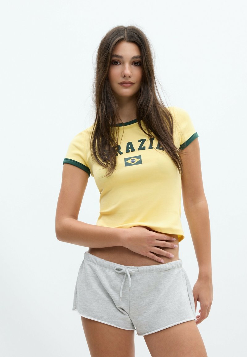 Young woman with long brown hair wearing a yellow Brazil t-shirt and gray shorts, standing with one hand on her stomach.