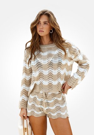 Mujer con jersey de punto beige y blanco con patrón de ondas y pantalones cortos a juego, sosteniendo un bolso beige con textura, de pie sobre fondo blanco.
