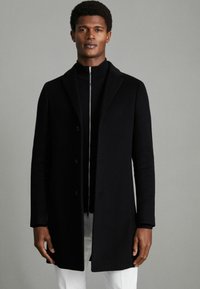 Cappotto nero con design su misura, caratterizzato da un colletto a punta, chiusura con bottoni e texture liscia, indossato sopra un maglione nero con zip.