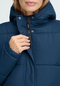 Temno modra puffer jakna z zadrgo spredaj, nastavljivo kapuco in zaponkami. Gladka tekstura z vidnim šivnim vzorcem.