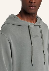 Olivgrön hoodie med dragsko, ribbade ärmmuddar och tryckt logga på bröstet. Tyget är strukturerat och något löst sittande.