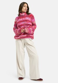 Pinker, grob gestrickter Pullover mit hohem Kragen und weiten Ärmeln, kombiniert mit cremefarbenen, weiten Hosen und burgunderfarbenen Schuhen.