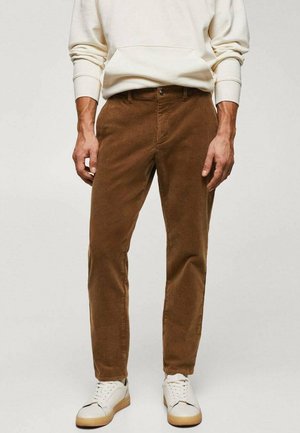 Mand iført brune fløjlsbukser kombineret med hvide sneakers og en cremefarvet sweatshirt, stående mod en ensfarvet baggrund.