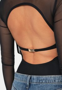 Μαύρο διάφανο bodysuit μακρυμάνικο με κοπή χαμηλά στην πλάτη, διαθέτοντας ένα κομψό χρυσό μεταλλικό κούμπωμα και μια ριγέ μαύρη ταινία. Συνδυασμένο με μπλε τζιν.