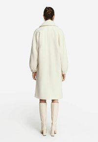 Krämliknande ullkappa med hög krage, sänkta axlar och långa, voluminösa ärmar. Till plagget är det matchat med knähöga beige stövlar.