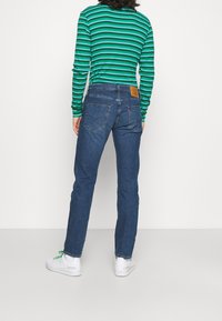 Jeans de mezclilla azul con corte recto, con una parche de cuero marrón en la cintura, combinados con una camisa de manga larga de rayas verdes y azules.