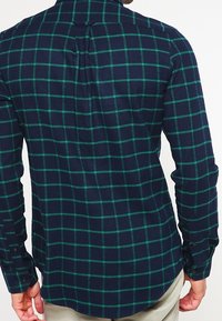 Camicia di flanella blu navy con motivo a quadri verdi. Presenta un colletto button-down e maniche lunghe. Vestibilità aderente con orlo curvo.