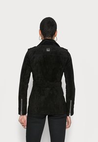 Czarna zamszowa kurtka z paskiem w talii, zapinanymi mankietami na zamek i kołnierzem. Posiada kwadratową łatkę z logo na plecach i dopasowany krój.