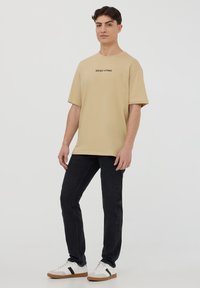 T-shirt di cotone beige a maniche corte e scollo rotondo, con la scritta "WEST COAST" in nero. Abbinata a jeans scuri e sneaker.