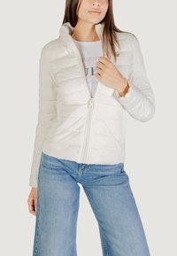 Witte pufferjack met een hoge kraag, ritssluiting aan de voorkant en doorgestikte textuur. Draag het over een lichtgrijs T-shirt en combineer het met blauwe wijde jeans.