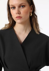 Abito avvolgente nero in tessuto morbido con scollo a V profondo e maniche larghe. La modella indossa orecchini a cerchio dorati, che evidenziano un design minimalista.