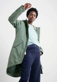 Zelená parka s texturovaným povrchem, se zipem a knoflíkovým zapínáním, huňatým límcem a bočními kapsami. Nosená přes pruhované tričko a manšestrové kalhoty.