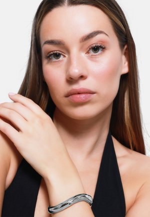 Metallarmband mit einem glatten, gewellten Design in einem glänzenden Silber-Finish, getragen am Handgelenk einer Person mit langen braunen Haaren.
