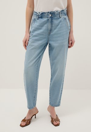 Persona con vaqueros de tiro alto azul claro con cintura fruncida y sandalias marrones de tacón abierto, de pie frente a un fondo liso.