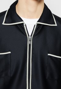 Närbild av svart jacka med vitt kantband på kragen, dragkedjan och fickkanterna, bärs över en vit rundhalsad tröja.