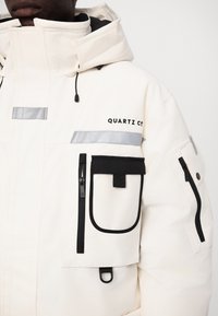 Bijela izolirana jakna s kapuljačom, sivo-crnim naglascima, višestrukim patentnim zatvaračima i logotipom "QUARTZ Co." na gornjoj lijevoj strani.