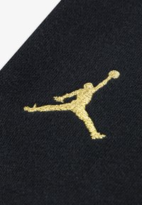 Černá tkanina s texturovaným povrchem, na níž se nachází výrazné zlatě vyšívané logo Jumpman v dynamické póze, které předvádí atletický design.